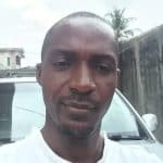 A portrait of a young man with a shaved head, standing outdoors near a car, with a blurred background of buildings and trees. Focused on natural, casual photography for personal or lifestyle branding.